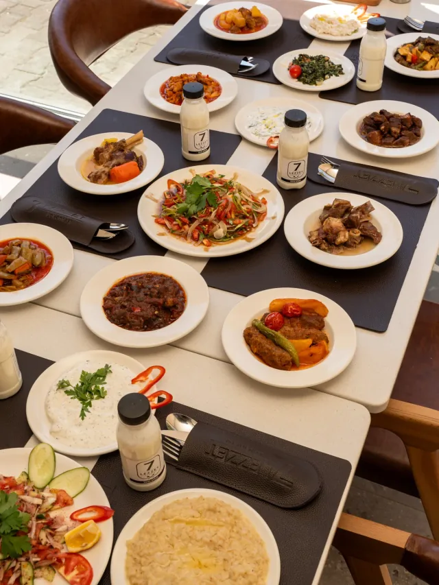 7 Lezzet Lokantası'nda kurulmuş sofra: tencere yemekleri, köfte, salata ve cacık; markalı ayran şişeleri ve deri amerikan servisler.